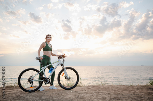 Beautiful young brunette woman resting after bicycle ride at sunrise. Model listening to music with wireless earbuds in the morning.