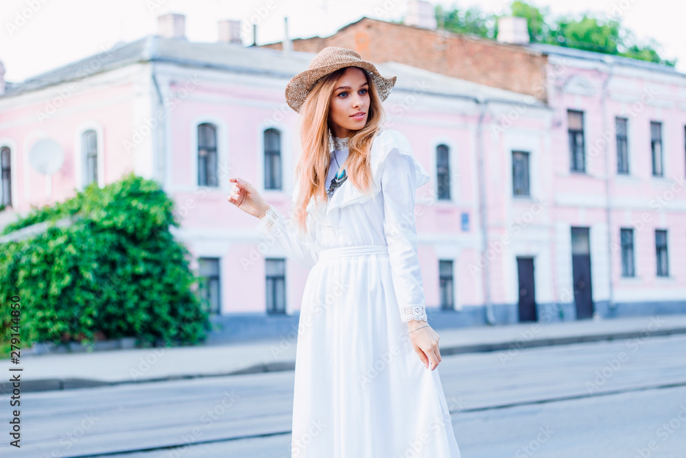 Girl traveling in vintage white dress with hat stands near the road amid the pink house near the road