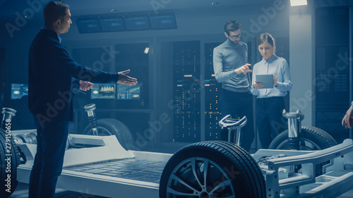Automobile Engineers Discussing and Designing Electric Car Chassis Platform, Using Tablet Computers with 3D CAD Software. In Automotive Innovation Facility Vehicle Frame with Wheels Engine and Battery © Gorodenkoff