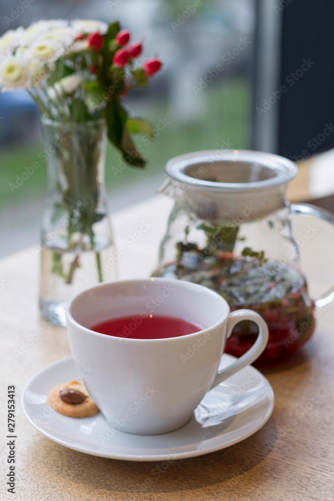 Cup of red herbal tea, cookie, teapot, vase with flowers on the table. Close-up, copy space, vertical format Cup of red herbal tea, cookie, teapot, vase with flowers on the table. Close-up, copy space, vertical format