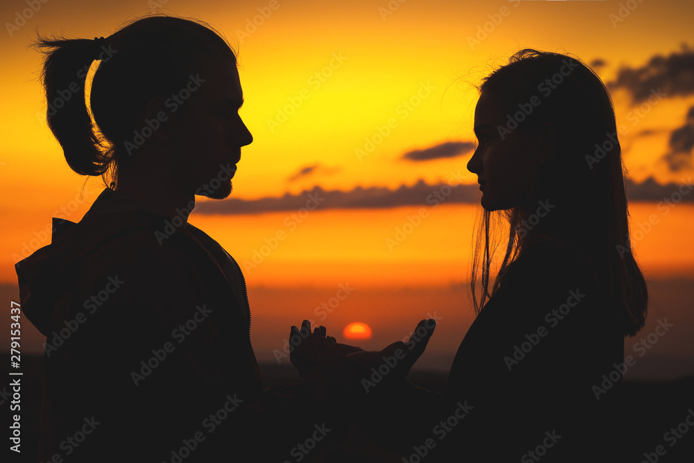 Close-up of the silhouettes of a young millenial couple in love hands are holding the sun with a man and girl against the sunset golden sky. They look at each other. Young family