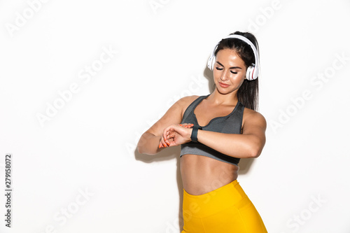 Smiling pleased beautiful young sports fitness woman isolated over white wall background using watch clock listening music with headphones.