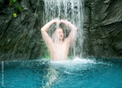 young man enjoys the falling water under the splashing waterfall in the turquoise spa pool