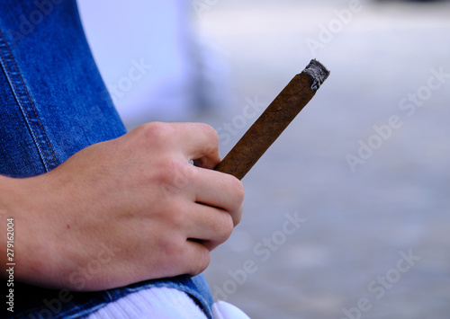 Hand of a young white male holding a lit up cigar while resting hand of jeans.