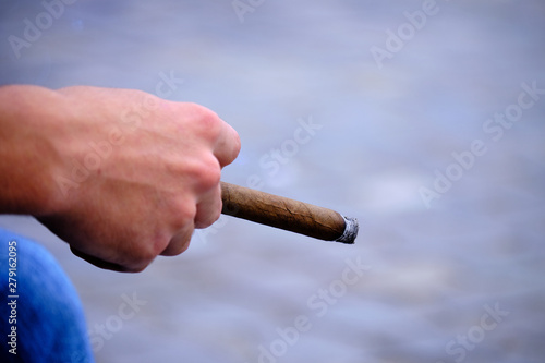 Hand of a young white male holding a lit up cigar while resting hand of jeans.