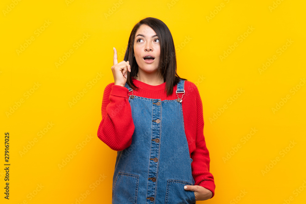 Young Mexican woman with overalls over yellow wall thinking an idea pointing the finger up Young Mexican woman with overalls over yellow wall thinking an idea pointing the finger up