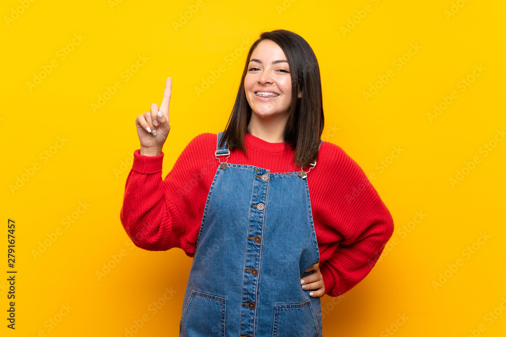 Young Mexican woman with overalls over yellow wall showing and lifting a finger in sign of the best Young Mexican woman with overalls over yellow wall showing and lifting a finger in sign of the best