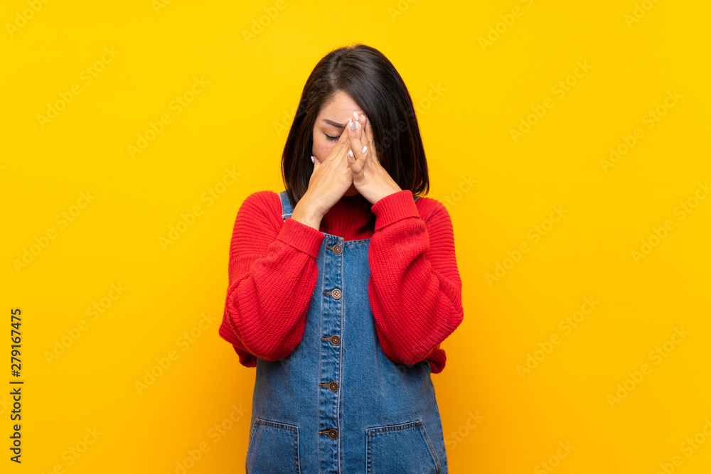 Young Mexican woman with overalls over yellow wall with tired and sick expression Young Mexican woman with overalls over yellow wall with tired and sick expression
