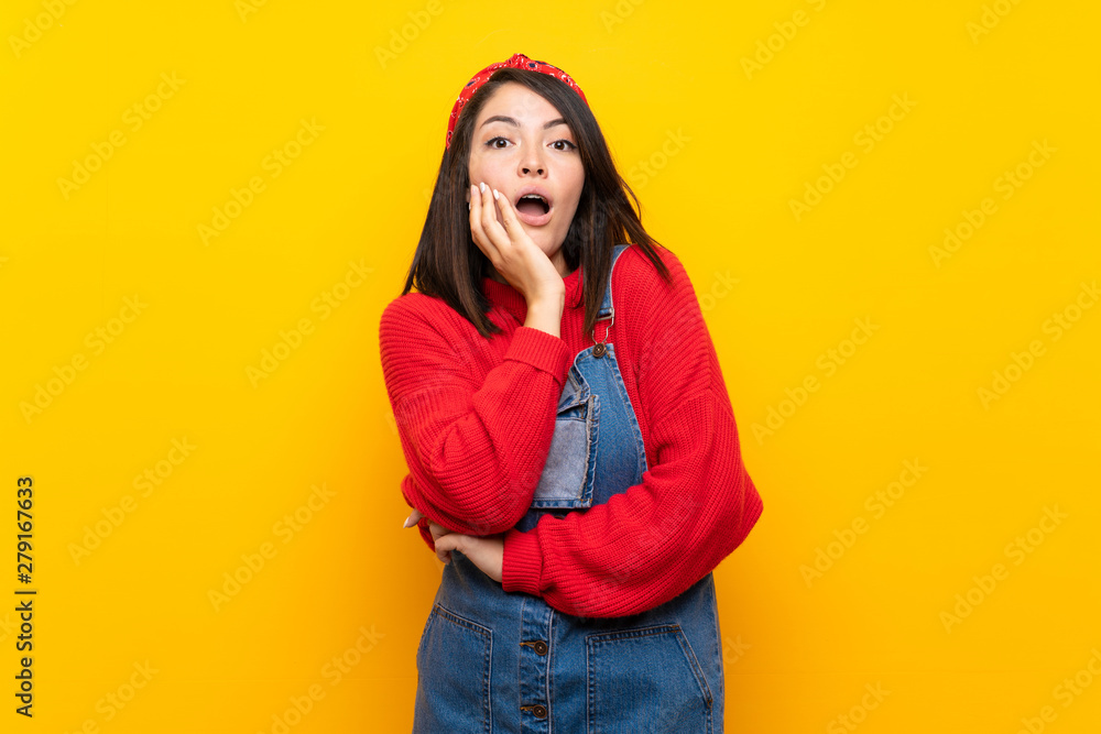 Young Mexican woman with overalls over yellow wall surprised and shocked while looking right Young Mexican woman with overalls over yellow wall surprised and shocked while looking right