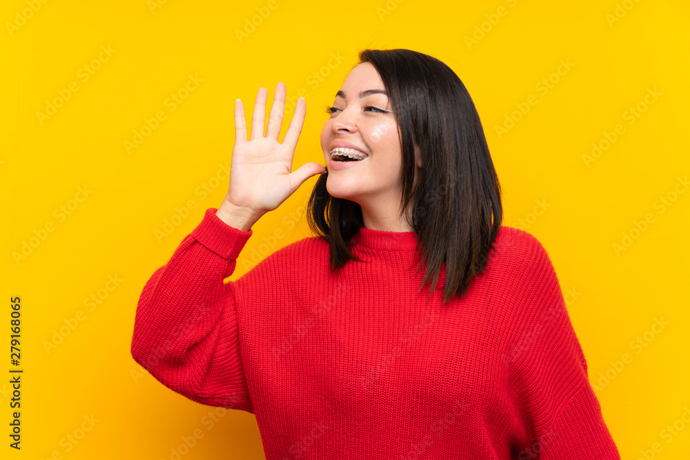 Young Mexican woman with red sweater over yellow wall shouting with mouth wide open Young Mexican woman with red sweater over yellow wall shouting with mouth wide open