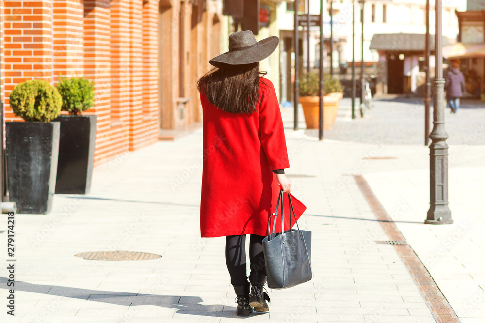 Stylish beautiful woman walking in street. Girl wearing red coat, black hat and holding trendy bag. Fashion outfit, autumn trend, accessories. Women fashion, urban style