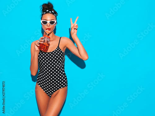 Young beautiful sexy smiling hipster woman in sunglasses.Girl in summer peas swimwear bathing suit.Posing near blue wall,drinking fresh cocktail smoozy drink.Shows peace sign