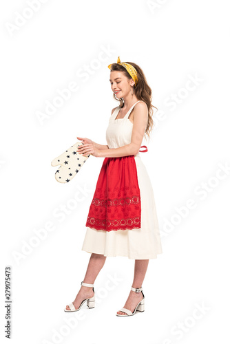 full length view of young happy housewife in dress and apron holding oven mitten isolated on white