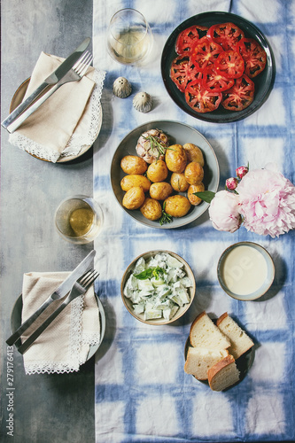 Summer vegetarian dinner table for family or friends. Young baked potatoes, tomato carpaccio, cucumber salad, bread, wine, sauce and flowers over linen tablecloth. Flat lay, space