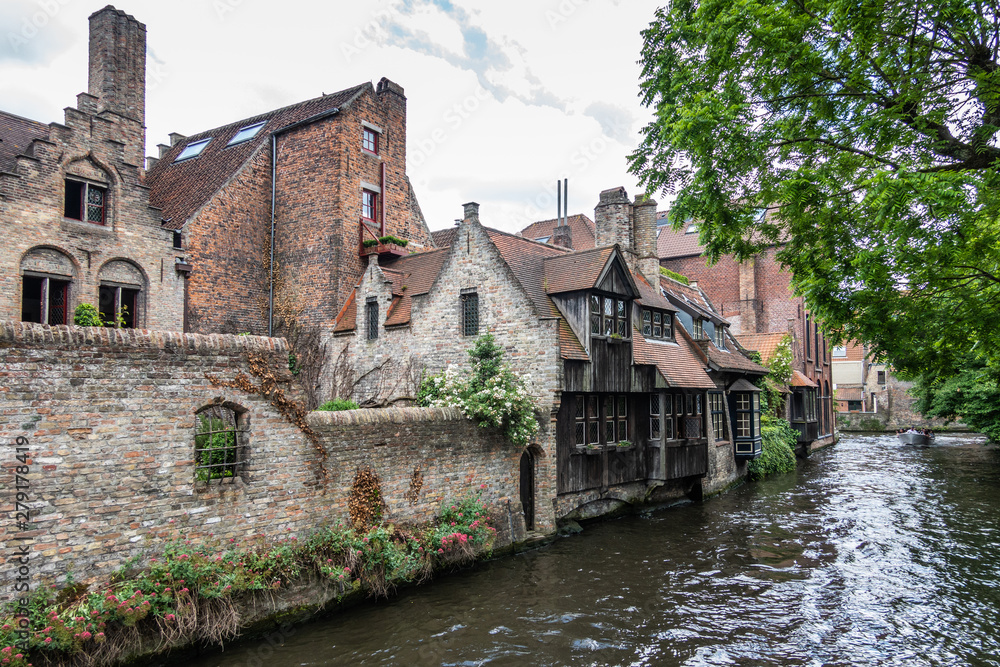 Bruges, Flanders, Belgium - June 17, 2019: Dark brown wooden facade among other brown brick stone facades along black water canal. Green foliage and white clouds. Bruges, Flanders, Belgium - June 17, 2019: Dark brown wooden facade among other brown brick stone facades along black water canal. Green foliage and white clouds.