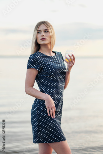 A young attractive blond woman, in a blue dress, walks along the seashore. Portrait of a smiling woman on vacation with a glass of wine.