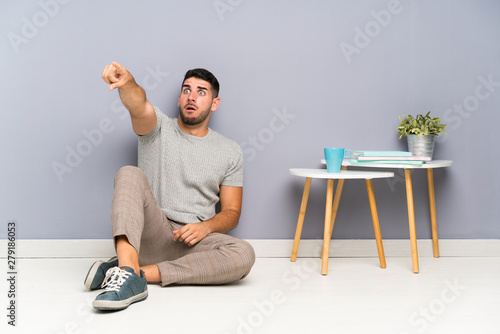 Young handsome man sitting on the floor pointing away