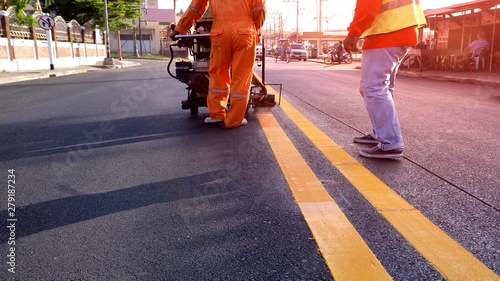 Selective focus of road workers with thermoplastic spray road marking machine working to paint yellow line on asphalt road surface with flare light in evening time, construction concept