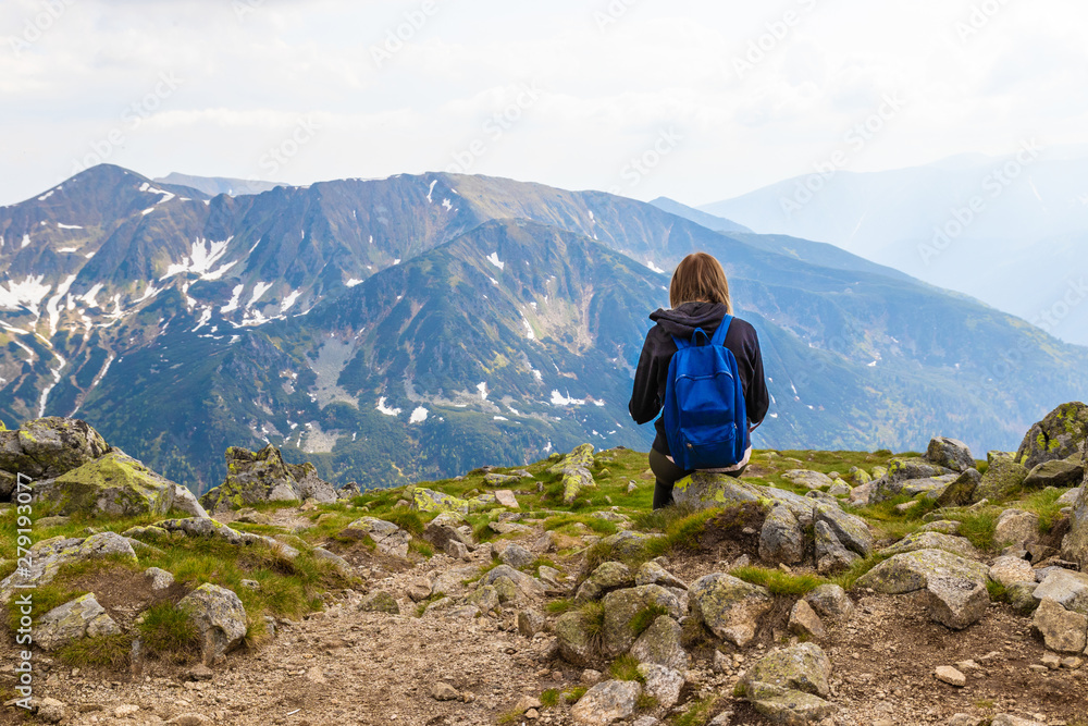 A young blonde travels with a blue backpack, sits on top of a mountain and takes pictures of the green mountain scenery. Back view