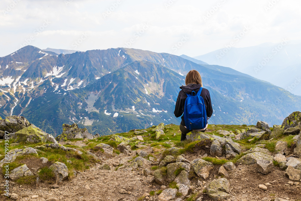Fototapeta premium A young blonde travels with a blue backpack, sits on top of a mountain and takes pictures of the green mountain scenery. Back view