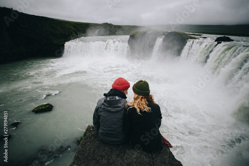 Vászonkép Couple of travellers in winter warm clothing sit on steep dangerous cliff next to waterfall, watch falling water, calm and astonished by power of nature