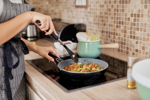 Charming Caucasain brunette in apron standing next to stove in kitchen and cooking healthy dinner. photo