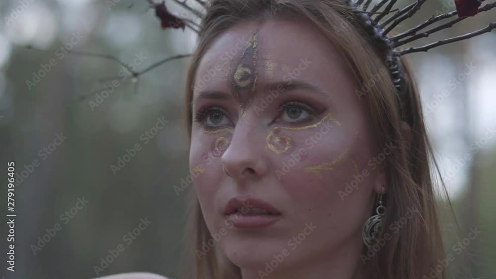 Portrait of an attractive dryad or forest fairy with a wreath of branches on the head and painted the third eye on the forehead dancing under the trees. Ancient ritual of forest creature. Slow motion. Portrait of an attractive dryad or forest fairy with a wreath of branches on the head and painted the third eye on the forehead dancing under the trees. Ancient ritual of forest creature. Slow motion.