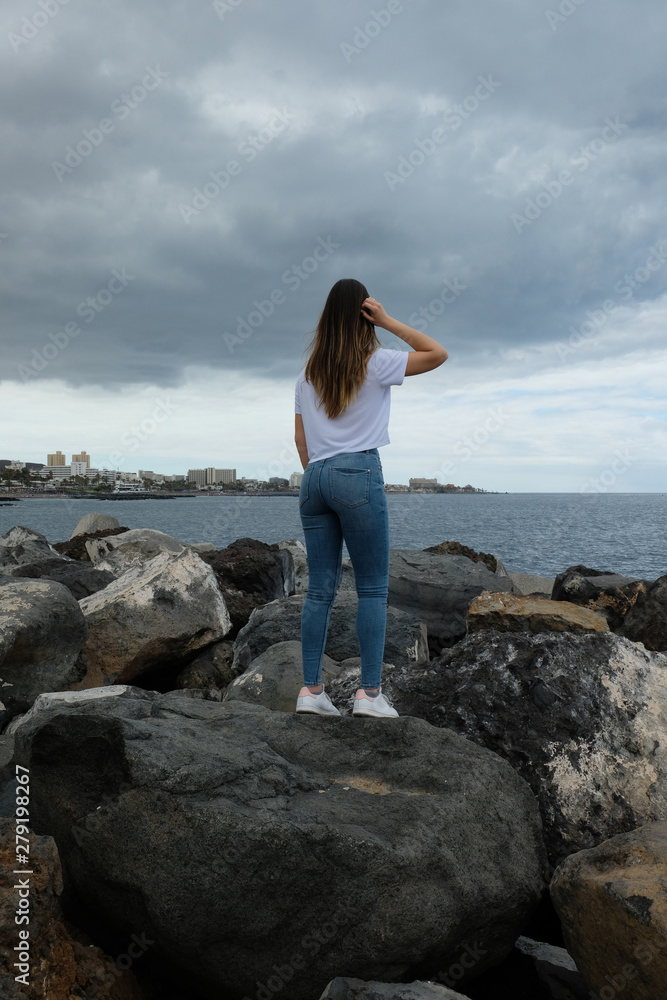 Beautiful girl standing on beach rocks looking to the sea