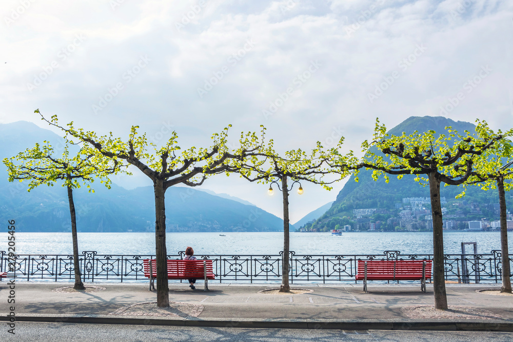 Woman rests on bench at the promenade of luxury resort in Lugano and admires the beautiful scenery of Lake Lugano, surrounded by Alps mountains on spring morning, the canton of Ticino, Switzerland.