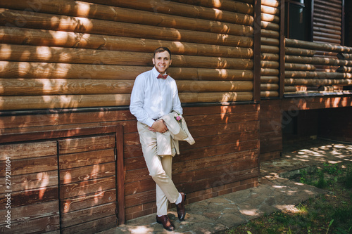 Stylish groom stands near the wooden house in the park, in a beige suit and smiles. Morning groom. Wedding portrait of a young, handsome man. Photography and concept.