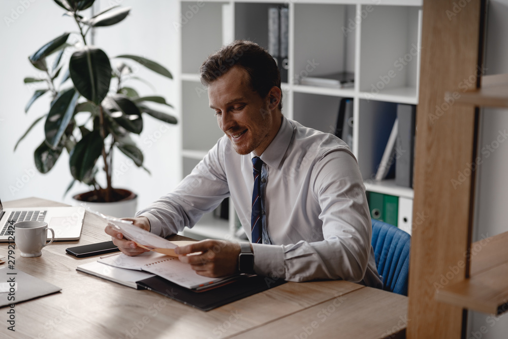 Happy young entrepreneur sitting with envelope in his hands and smiling while getting new documents Happy young entrepreneur sitting with envelope in his hands and smiling while getting new documents
