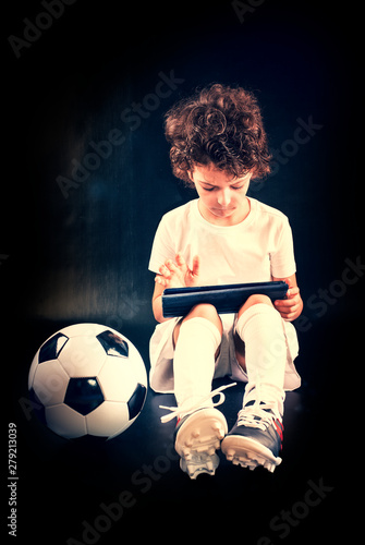 Puzzled little boy soccer player with soccer ball, using tablet wall computer isolated on black wall background. People emotions, sport family leisure lifestyle concept © kravik93