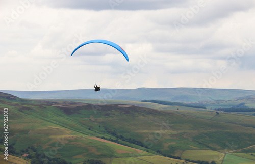 Blue paraglider flying above valley with green mountain and clouds in the background