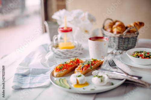 norway breakfast. Toasts with salmon and cream-cheese, boiled eggs served on a white plate standing on a white wooden table with vegetable salad, coffee, jar of orange juice and croissants
