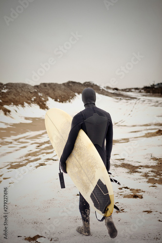 Canvas Print Rear view of man wearing wetsuit walking across along snowy beach carrying surfb