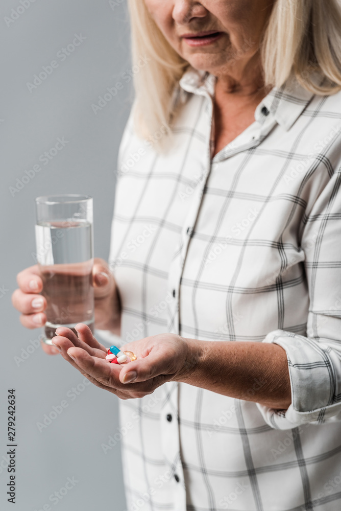 cropped view of senior woman holding pills and glass of water isolated on grey cropped view of senior woman holding pills and glass of water isolated on grey