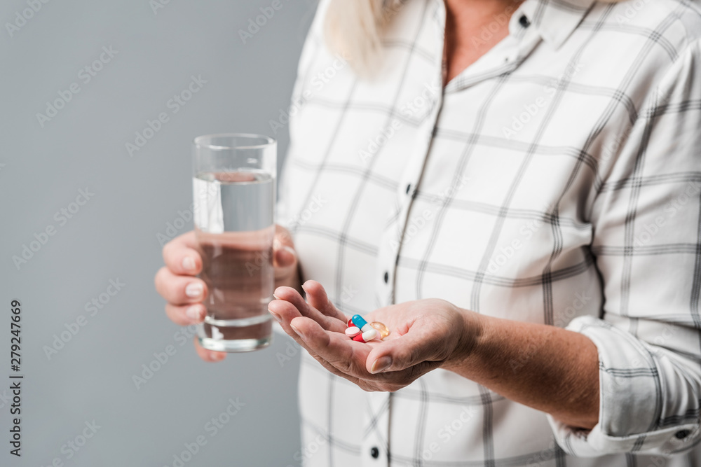 cropped view of retired woman holding pills and glass of water isolated on grey cropped view of retired woman holding pills and glass of water isolated on grey