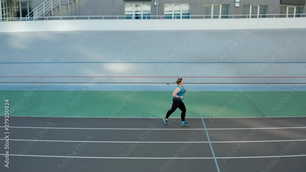 Aerial side view of plus size female in sportswear jogging at stadium on sunny morning. Active fat woman runner exercising on city sports ground, intense cardio training, effective weight loss concept Aerial side view of plus size female in sportswear jogging at stadium on sunny morning. Active fat woman runner exercising on city sports ground, intense cardio training, effective weight loss concept