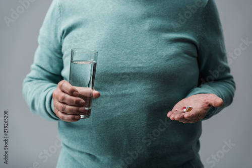cropped view of retired man holding pills and glass of water isolated on grey