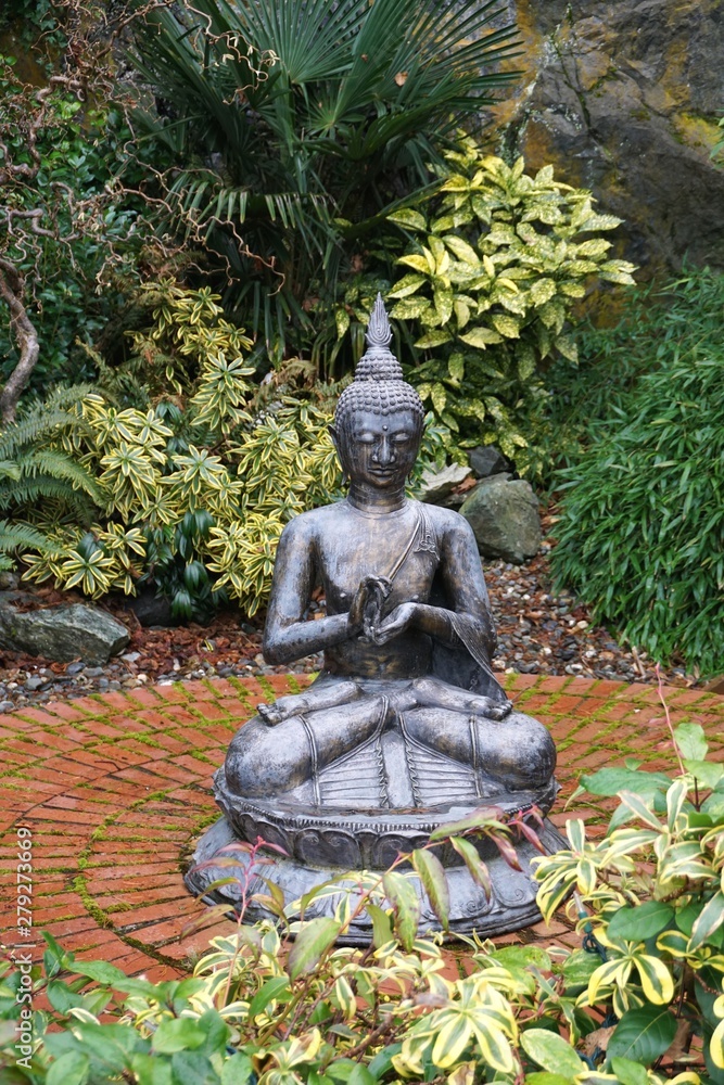 Antique Buddha statue on brick patio surrounded by lush green foliage in secret meditation garden Antique Buddha statue on brick patio surrounded by lush green foliage in secret meditation garden