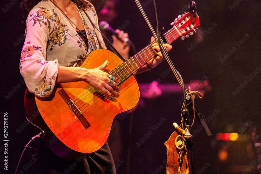 Obraz premium Band member uses guitar and mic stand. A close up view on a stylish lady band member playing a guitar during a live music set. Blurred musicians are seen in the background. Details on stage at gig.