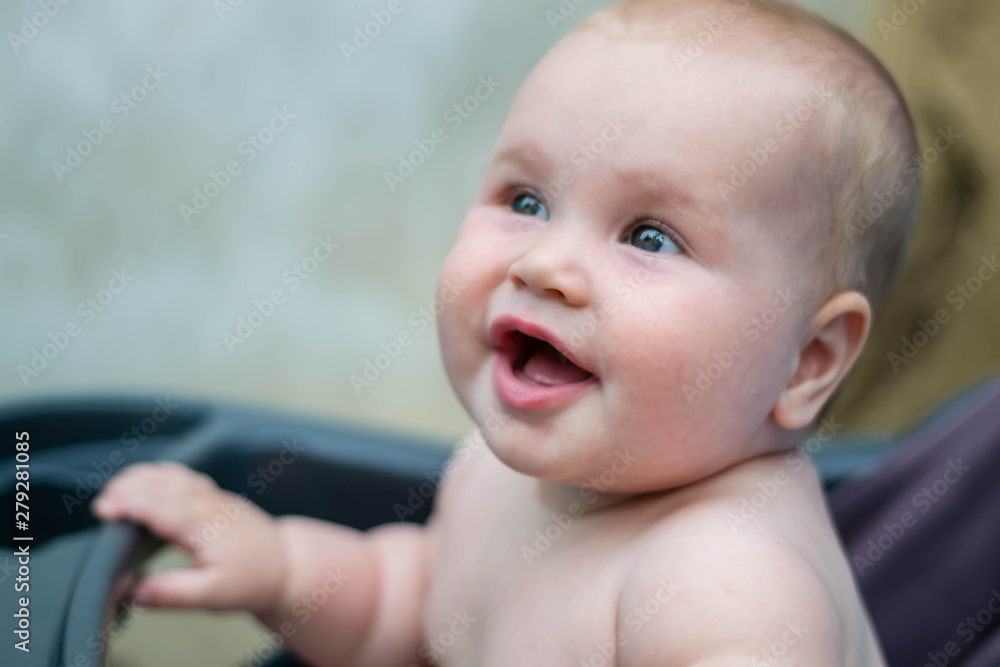 Little cute baby girl is sitting in the carriage and smiles. A child of the Caucasian appearance, undressed. The girl laughs. Summer garden, green trees, heat. Little cute baby girl is sitting in the carriage and smiles. A child of the Caucasian appearance, undressed. The girl laughs. Summer garden, green trees, heat.