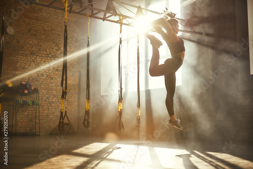 Perfect flexibility. Full length of athletic woman in black sport clothing jumping while exercising in professional semi-dark gym.