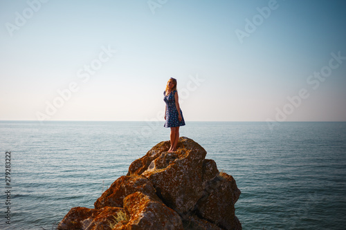 A girl in a dress stands on a stone and looks at the blue sea