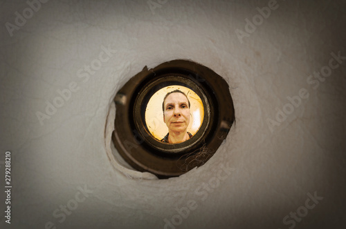 Woman seen out of focus through the old dirty peephole of the front door of a dark apartment