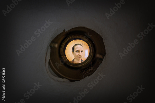 Woman seen out of focus through the old dirty peephole of the front door of a dark apartment