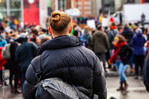 Activists march against global warming. A young millennial man is seen from behind, with a hair bun and backpack, watching blurry eco-activists march against climate change in the background