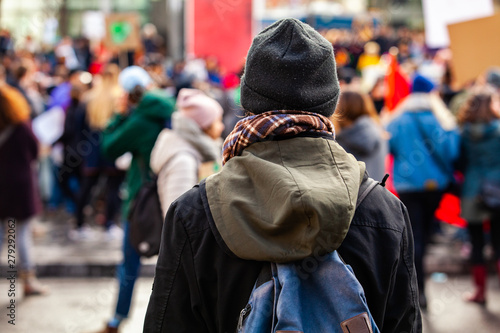 Person watches ecological rally. A young make is viewed from the back, wearing a hat and backpack, contemplating activists stage an environmental demonstration in a blurred background