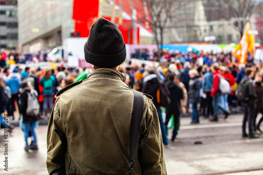 Man watches people march for environment. A young man is seen from the back, wearing an olive jacket and wooly hat, as eco-activists march against global warming in a blurry background. Man watches people march for environment. A young man is seen from the back, wearing an olive jacket and wooly hat, as eco-activists march against global warming in a blurry background.