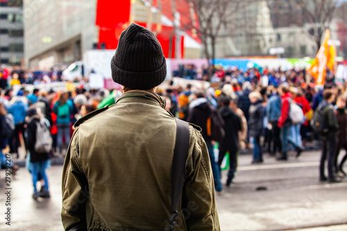 Man watches people march for environment. A young man is seen from the back, wearing an olive jacket and wooly hat, as eco-activists march against global warming in a blurry background.